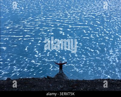 Vue aérienne d'une personne en veste rouge debout sur un éperon rocheux surplombant un vaste lac bleu gelé avec des textures de glace complexes au Népal. Banque D'Images