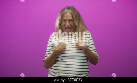 Femme blonde senior portant une chemise rayée pointe le doigt à la caméra avec la bouche ouverte et geste visible de la main en studio avec fond magenta ; ludique. Banque D'Images