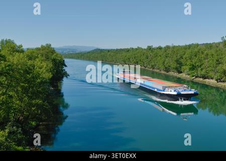 Vue en grand angle d'une grande barge de fret chargée de conteneurs maritimes naviguant sur une rivière calme entourée de forêts verdoyantes en France. Banque D'Images