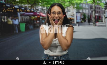 Jeune femme presse les mains sur les joues dans la surprise choquée dans la rue avec scène de café et lumières extérieures ; joie spontanée. Banque D'Images