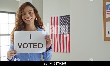 Femme latine souriante tient un panneau i voté avec les deux mains à l'intérieur d'un bâtiment de bureau de vote avec un drapeau américain visible ; fierté civique. Banque D'Images