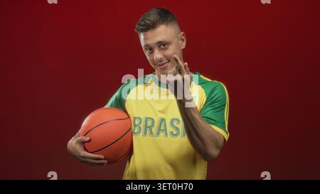 L'homme tient le basket-ball et fait des gestes de main de coeur en studio, portant le maillot brésilien ; fierté de l'esprit d'équipe. Banque D'Images