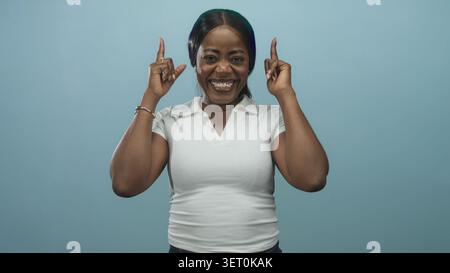 Femme en polo blanc pointe les index vers le haut et sourit largement montrant les dents en studio avec fond turquoise ; célébration de joie. Banque D'Images