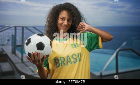 Femme tenant le ballon de football et donnant les pouces vers le bas sur la terrasse de la piscine portant le maillot du brésil et souriant ; fierté. Banque D'Images