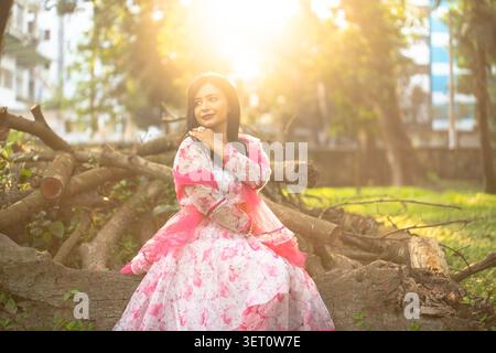 Une femme vêtue d'une robe à motifs floraux roses et blancs est assise en plein air contre une clôture en bois rustique pendant la lumière dorée du crépuscule. Banque D'Images