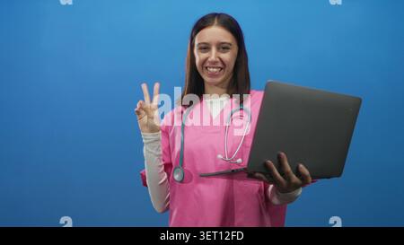 Jeune infirmière hispanique dans des gommages roses avec stéthoscope tient un ordinateur portable et fait signe de paix en studio ; télémédecine joyeuse. Banque D'Images