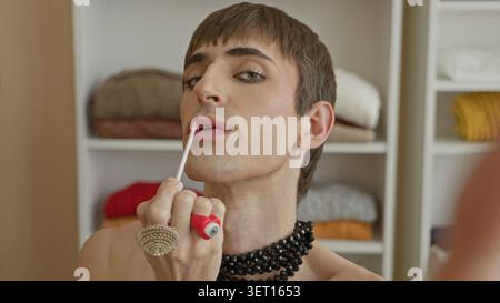 Jeune homme appliquant un gloss sur les lèvres avec une baguette, portant un collier de perles et des bagues tout en faisant face à un miroir et des étagères de garde-robe dans un studio ; identité conf Banque D'Images
