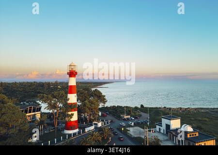 Vue aérienne Phare blanc rouge sur la côte de la mer Noire au coucher du soleil, Poti, Géorgie. Mer calme, nageurs, voitures, parc côtier, point de repère de navigation, maritime sa Banque D'Images
