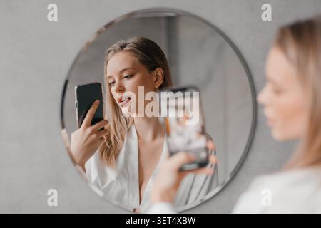 Jeune femme caucasienne prenant Selfie avec smartphone devant le miroir, réglage de la maison Banque D'Images