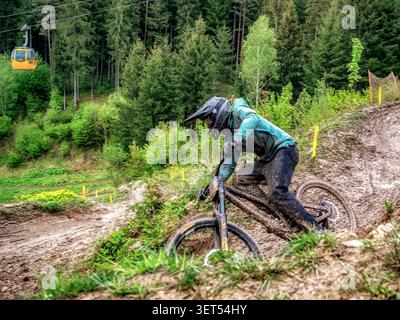 Ružomberok, Slovaquie - 17 mai 2026 : un coureur couvert de boue navigue dans un virage humide sous la ligne du téléphérique au Bikepark Malino Brdo après une forte pluie créa Banque D'Images