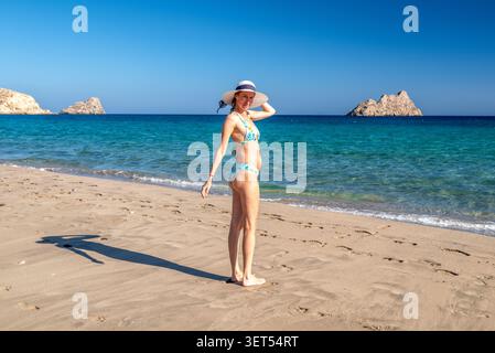 Jeune femme en bikini et chapeau de soleil debout sur la plage de sable Mazida Ammos en Crète, Grèce, avec la mer turquoise, les îlots rocheux et le ciel bleu clair. Banque D'Images