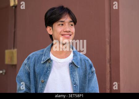 Portrait horizontal moyen d'un jeune homme joyeux souriant d'Asie du Sud-est Thai Isan dans une veste en denim regardant loin tout en se tenant debout contre un brun Uni Banque D'Images