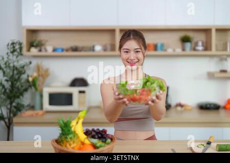 Une jeune femme joyeuse en vêtements d'action présente un bol de salade fraîche dans sa cuisine, mettant l'accent sur une alimentation saine et une vie vibrante. Banque D'Images