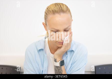 Femme souffrant de maux de dents sévères ou de douleurs à la mâchoire, tenant un mouchoir sur son visage, assise dans une salle d'attente de clinique dentaire avant un rendez-vous, Needin Banque D'Images