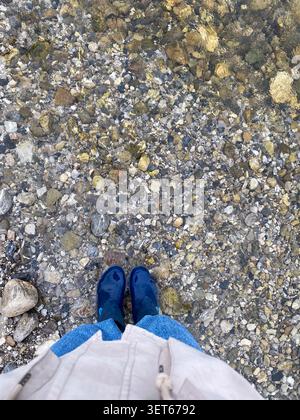 Personne portant des bottes de pluie bleues debout dans un ruisseau peu profond avec un fond de galet Banque D'Images
