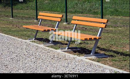 Une paire de bancs publics vides ornés de lattes en bois brun clair et de supports en acier gris se trouvent sur une parcelle d'herbe le long d'un chemin de galets bordant Banque D'Images