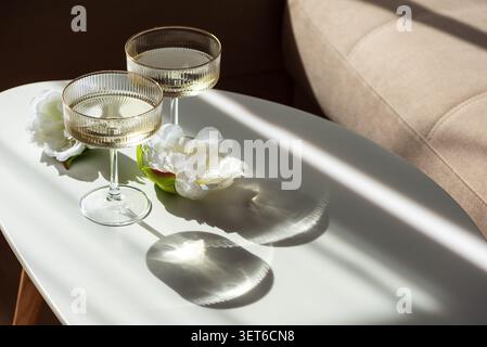 Deux verres coupés côtelés remplis de vin mousseux et de délicates fleurs blanches reposant sur une table blanche propre. Baigné de lumière du soleil créant capt Banque D'Images