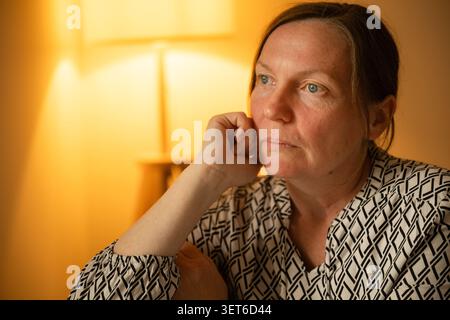 Femme d'âge moyen réfléchie reposant la main contre la bouche, regardant inquiet dans une lumière intérieure chaude, mise au point sélective Banque D'Images