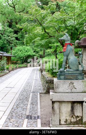 Statue de renard kitsune en bronze avec bavoir rouge à côté d'une allée de pierre au Fushimi Inari Taisha, Kyoto Japon, des érables verts remplissent l'arrière-plan du sanctuaire Banque D'Images