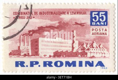ROUMANIE - 1963 avril 10 : timbre de la poste aérienne d'interdiction brun-rouge et bleu foncé 55 représentant le complexe industriel de transformation du bois à Târgu Jiu et la silhouette d'un avion. L'illustration montre une grande installation industrielle moderniste avec des blocs d'usine à fenêtres multiples proéminentes, plusieurs cheminées hautes dégageant une fumée épaisse, et des installations de traitement spécialisées, tandis qu'un avion de transport vole à travers le ciel nuageux au-dessus. Cette imagerie met en évidence l'industrialisation socialiste roumaine du milieu du XXe siècle, mettant l'accent sur la fabrication du bois et la croissance économique planifiée par l'État Banque D'Images