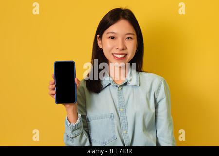 Femme asiatique souriante tenant smartphone avec écran vierge, fond jaune Banque D'Images