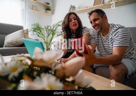 Couple heureux interagissant avec téléphone portable et ordinateur portable tout en se relaxant sur le canapé dans le salon Banque D'Images