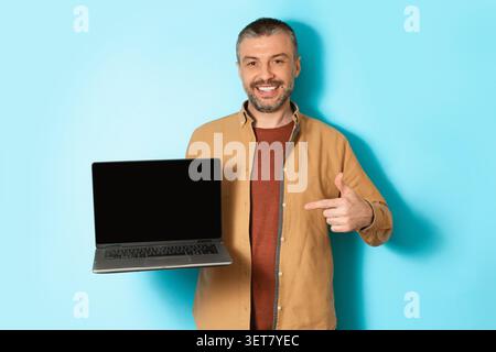 Homme souriant tenant l'ordinateur portable et pointant à l'écran, fond bleu, concept d'affaires Banque D'Images