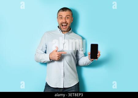 Homme caucasien excité affichant smartphone avec écran blanc, fond bleu, concept d'affaires Banque D'Images