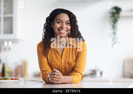 Souriant jeune femme afro-américaine se penchant sur le comptoir de cuisine avec salade saine, concept de style de vie Banque D'Images