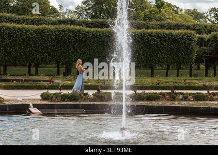 Une femme marche le long d'un chemin près d'une fontaine dans un jardin. Le soleil brille et les buissons verts sont tout autour. Le cadre est ouvert et animé avec de l'eau sp Banque D'Images