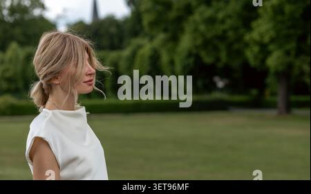Une femme marche dans un parc rempli d'arbres. Elle a les cheveux longs et porte une tenue blanche simple. C'est un après-midi chaud et il y a des gens dans le Banque D'Images