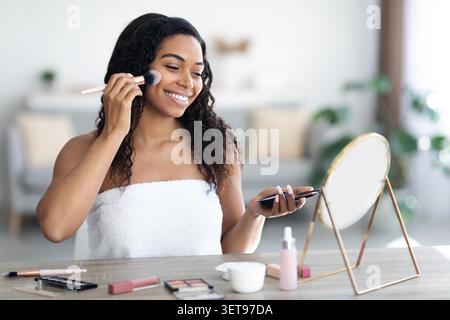 Femme noire heureuse appliquant le maquillage devant le miroir à la maison, concept de beauté Banque D'Images