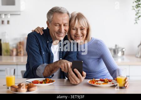 Heureux couple âgé caucasien assis à la table de cuisine, en utilisant smartphone ensemble Banque D'Images