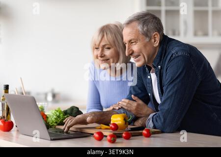 Heureux couple caucasien âgé cuisinant ensemble dans la cuisine, en utilisant l'ordinateur portable pour la recette, concept d'alimentation saine Banque D'Images