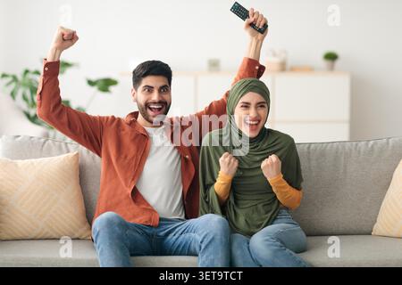 Excité jeune couple multiracial célébrant tout en regardant la télévision à la maison, heureux moment Banque D'Images