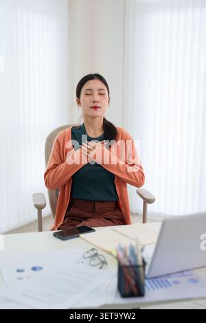 Femme d'affaires avec les mains sur la poitrine prenant une profonde respiration pendant la pause de travail pour la santé mentale et le soulagement du stress à l'espace de travail Banque D'Images