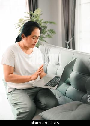 Femme asiatique assise sur un canapé à l'aide d'un ordinateur portable à la maison et levant la main, montrant le travail à distance, appel vidéo, réunion en ligne, communication numérique, domicile Banque D'Images