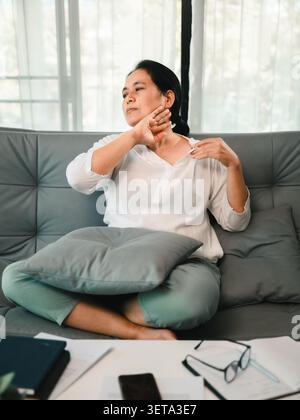 Femme asiatique assise sur un canapé à la maison avec un malaise au cou, touchant son épaule près de la paperasse et des lunettes, montrant la fatigue au bureau, une mauvaise posture, St Banque D'Images