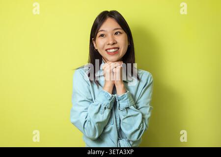 Sourire jeune femme asiatique avec les mains ensemble sur fond jaune, concept d'émotion heureuse Banque D'Images