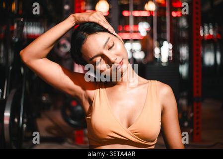 Gros plan d'une femme asiatique athlétique étirant son cou dans un gymnase sombre, les yeux fermés avec une main sur la tête, montrant le soulagement de tension, la mobilité, la récupération, min Banque D'Images