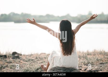 Vue arrière de la jeune femme asiatique dans une robe blanche assise au bord d'un lac paisible à bras ouverts, montrant la liberté, la libération émotionnelle, le bien-être de la nature, frais Banque D'Images