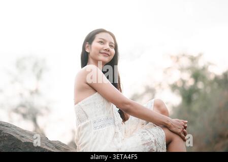 Jeune femme asiatique dans une robe de dentelle blanche assise sur un rocher et souriante dans une lumière naturelle douce, montrant un portrait extérieur paisible, un style de vie détendu, tra Banque D'Images