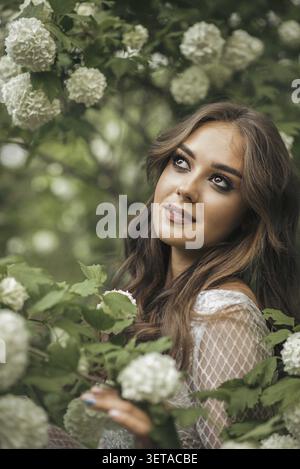 Portrait d'une belle fille dans une robe blanche avec de longs cheveux dans le jardin parmi les arbres à fleurs. sur les branches se trouvent de grandes fleurs blanches. la fille l'est Banque D'Images