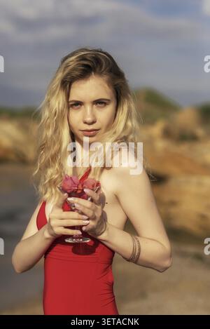 Portrait de Blonde en maillot de bain rouge sur Sandy Beach, elle tient du verre avec des fleurs dans ses mains profitant du soleil lors d'une belle journée sur la plage. Jeune femme Banque D'Images