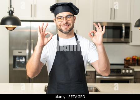 Chef cuisinier avec signe OK, nourriture délicieuse. Chef masculin millennial en uniforme de chefs. Chef homme cuisinant sur la cuisine. Chef cuisinier en chapeau et tablier. Banque D'Images