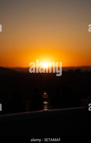 Bouteille d'eau en plastique sur une balustrade contre un coucher de soleil orange incandescent sur des collines lointaines, avec des silhouettes sombres et une ambiance de soirée calme. Banque D'Images