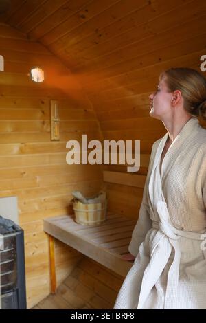 Femme dans un peignoir se relaxant dans un sauna traditionnel en bois, profitant d'une expérience de bien-être paisible et d'auto-soins. Banque D'Images