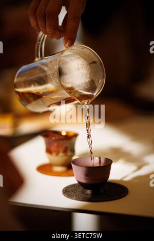 Une personne verse doucement du thé ambre foncé fraîchement infusé dans une tasse miniature faite à la main sur une table. L'arrière-plan montre une autre coulée de tasse de thé rustique molle Banque D'Images