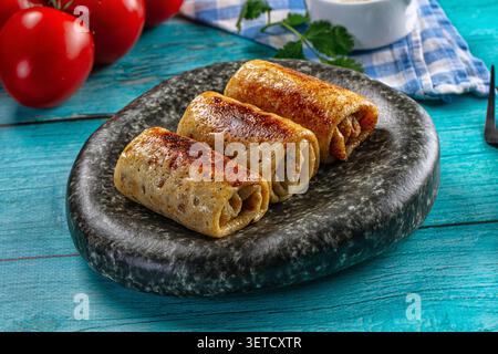 Crêpes frites brunes dorées avec garniture salée servies sur une assiette en pierre foncée. Fond en bois bleu rustique, tomates rouges fraîches, bol blanc, herbes vertes Banque D'Images