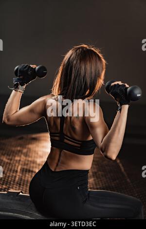 Fit femme dans les vêtements de sport effectue des exercices d'épaule avec des haltères, mettant en valeur le dos tonique et les bras. L'environnement moderne de gymnastique souligne son engagement envers un mode de vie sain et actif Banque D'Images